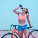 Jak przygotować organizm do sezonu na rower miejski znaczenie elektrolitów i witamin podawanych dożylnie Kobieta na rowerze w upale pije wodę podczas treningu w Dubaju
